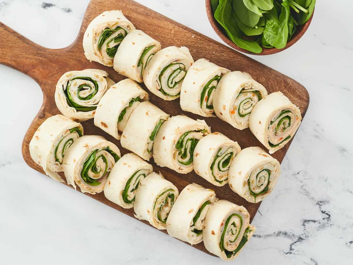 An overhead shot of 18 turkey pinwheels lined up into three rows on top of a wooden cutting board next to a wooden bowl of fresh spinach on a white marble background.